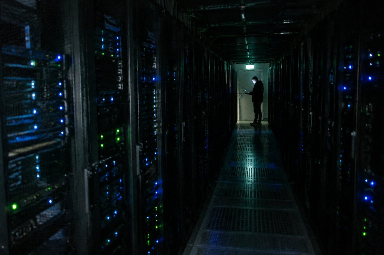 A dark data center hall: rows of server racks with blinking blue and green lights, one technician at the far end